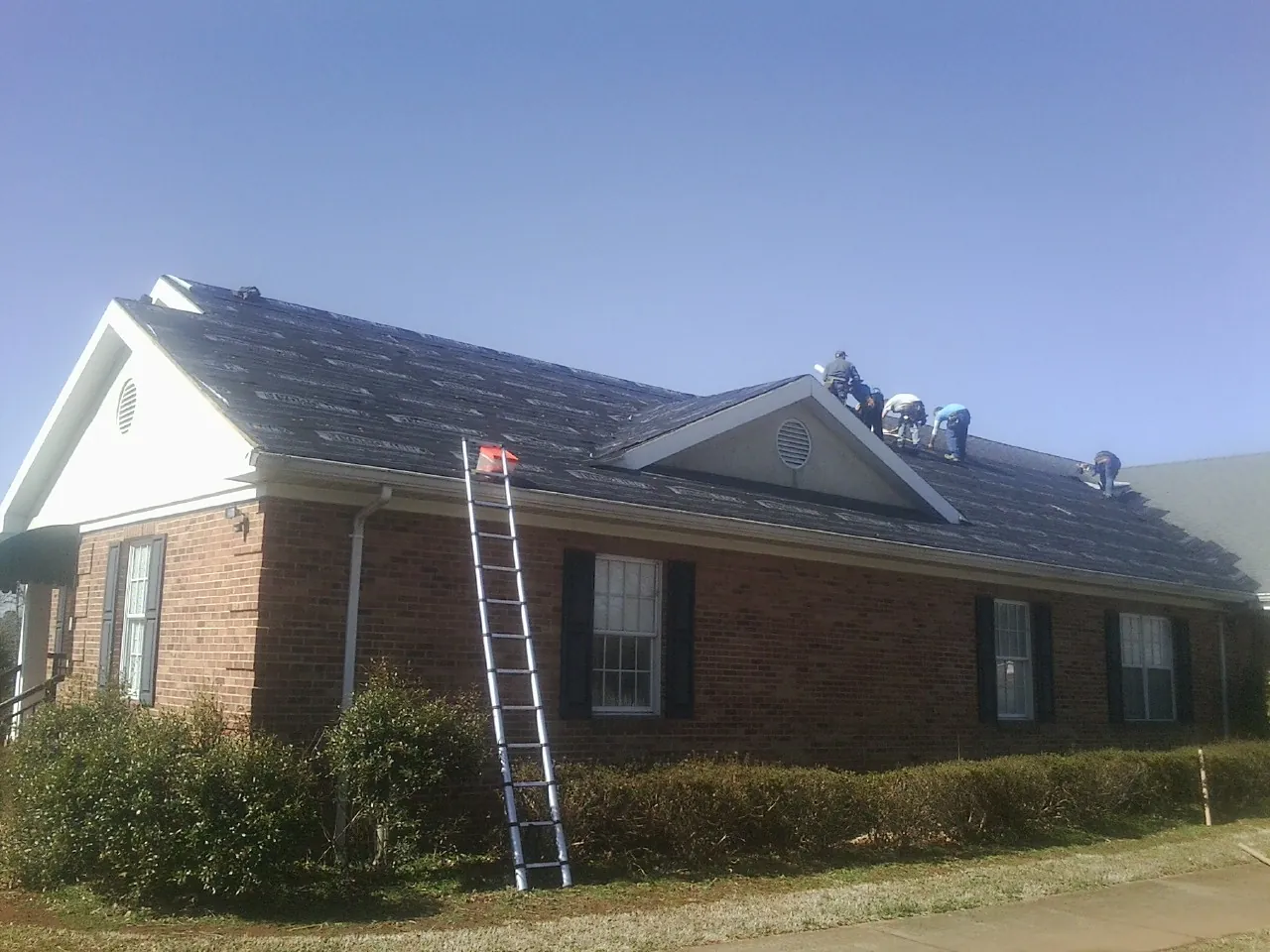 Crew tearing shingles off a brick ranch roof with a ladder set — Metal Roof Repair work by our crew in Sturtevant