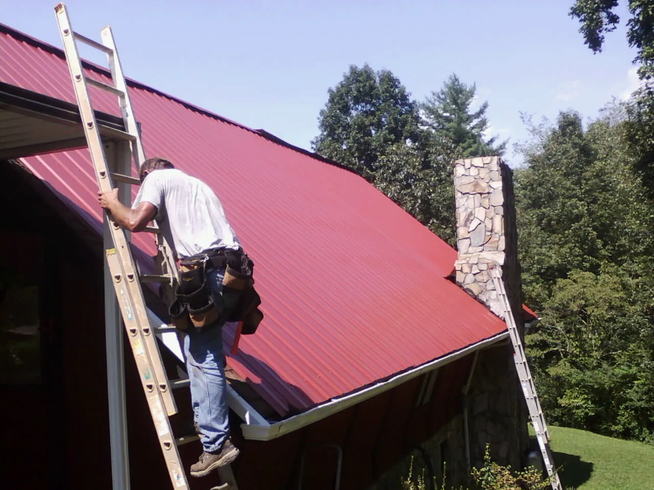 Roofer on a ladder beside a red metal roof and stone chimney — Soffit & Fascia Repair by Roofing Brothers in Sturtevant